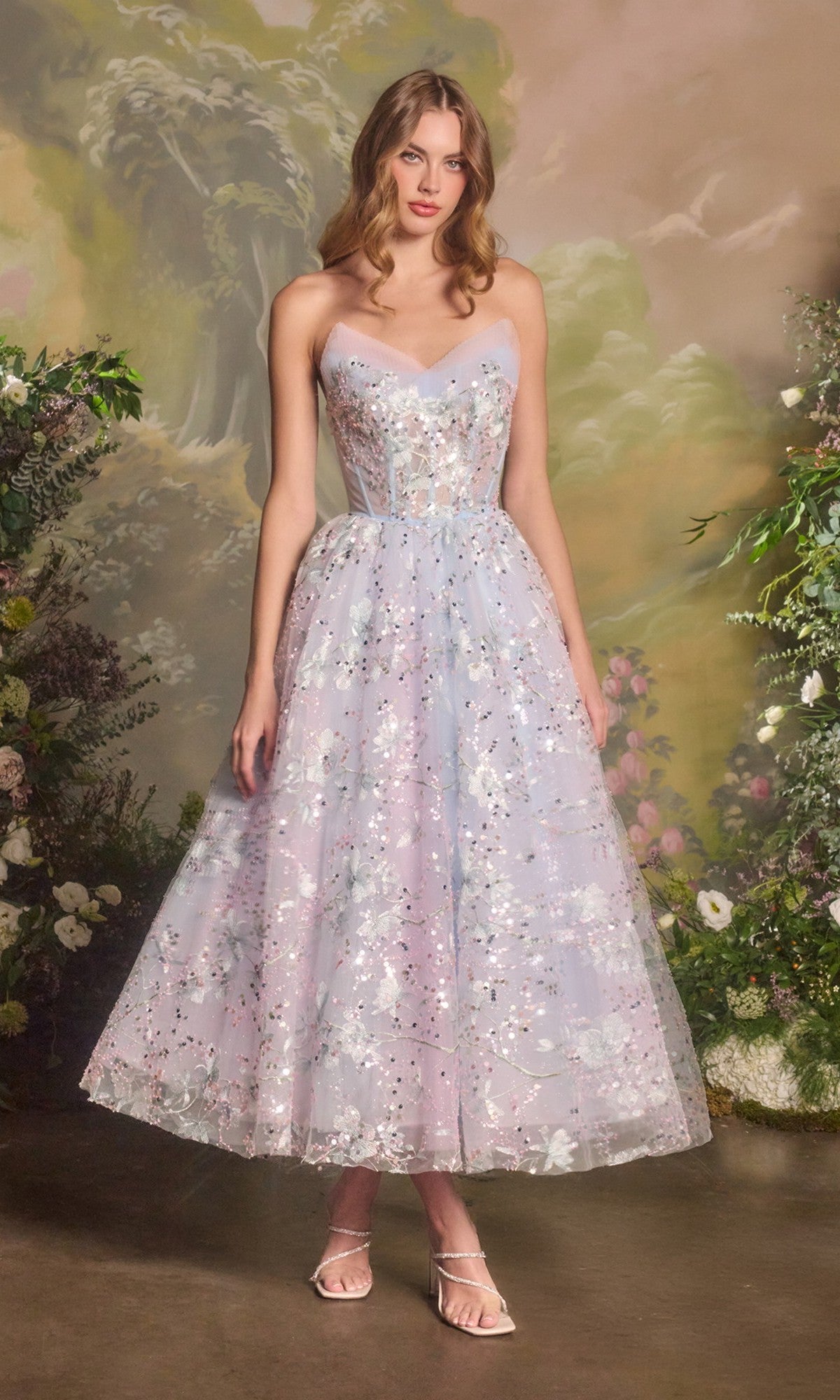 A woman stands indoors in the light blue Andrea & Leo A1485 strapless sequin prom dress, ankle-length with pastel floral patterns and metallic embellishments, surrounded by flowers and greenery.